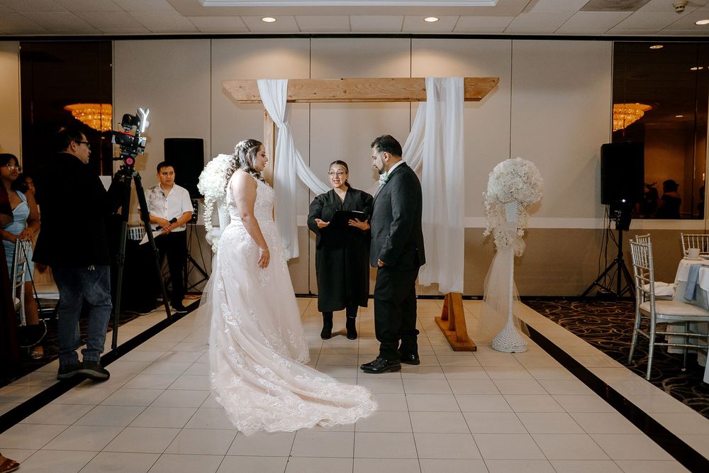 A wedding ceremony taking place in an indoor venue with white decorations and photographers capturing the moment.