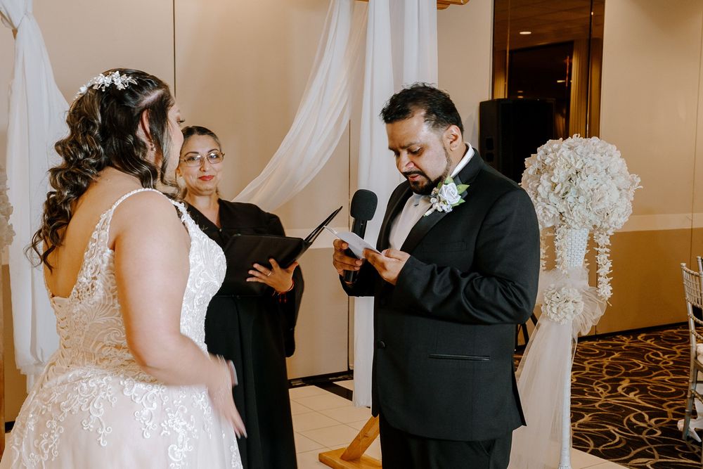 A couple exchanges vows during an indoor wedding ceremony with an officiant and white floral decorations.