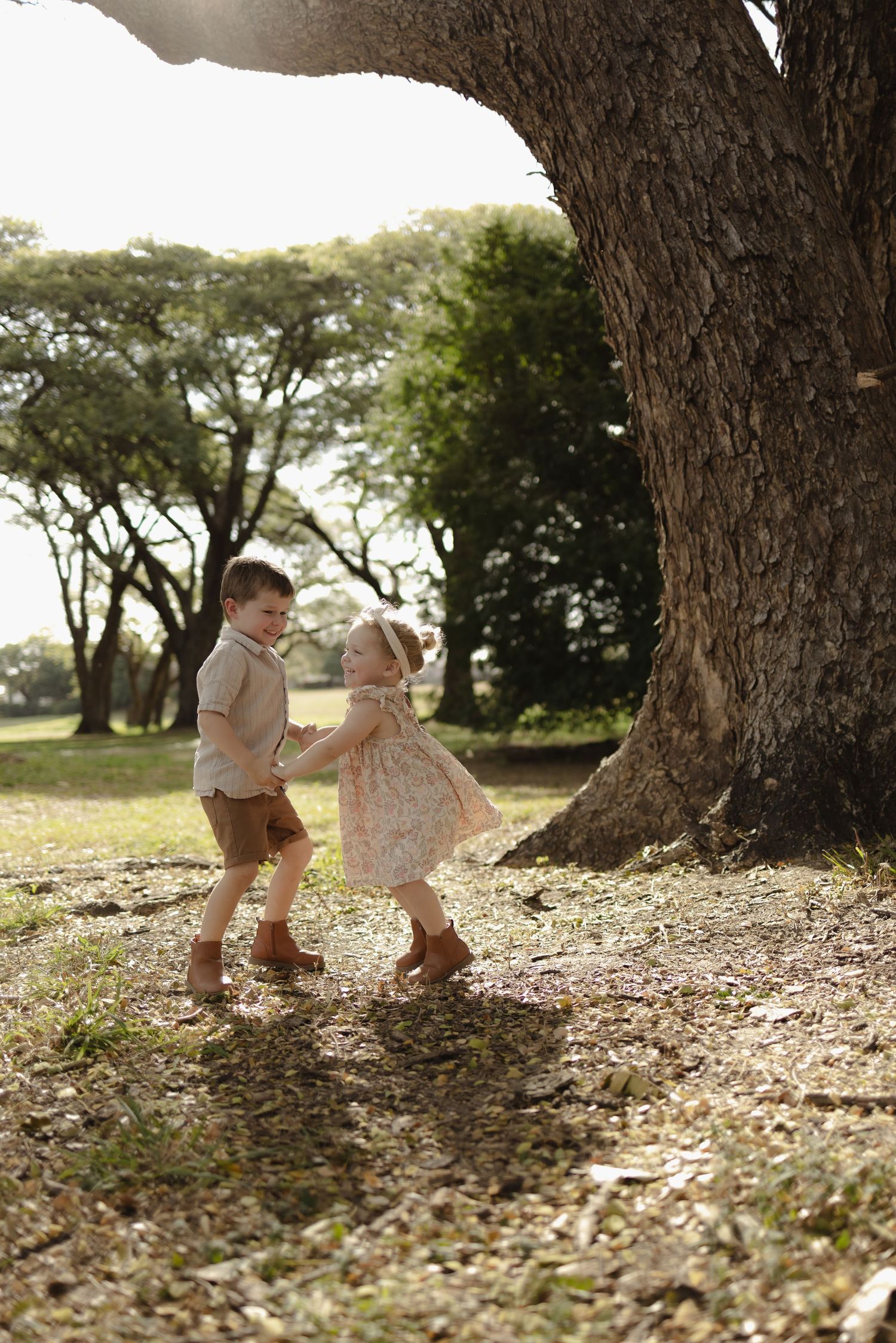 Children play together among fallen leaves near massive tree trunks in a park.