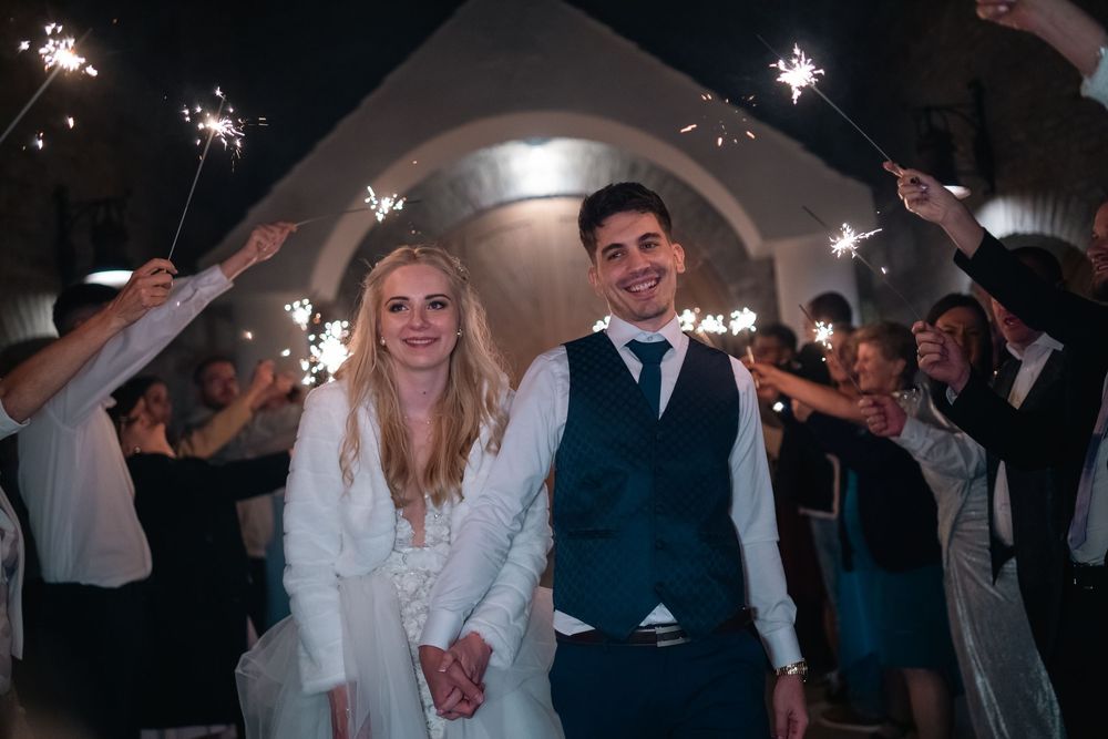 Wedding couple stands together under an archway while guests wave sparklers during their evening celebration exit.
