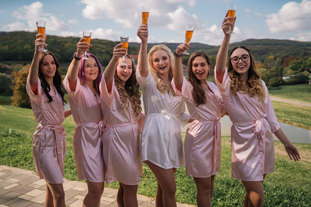 A group wearing matching pink satin robes toasts with champagne bottles while celebrating outdoors in a scenic countryside.