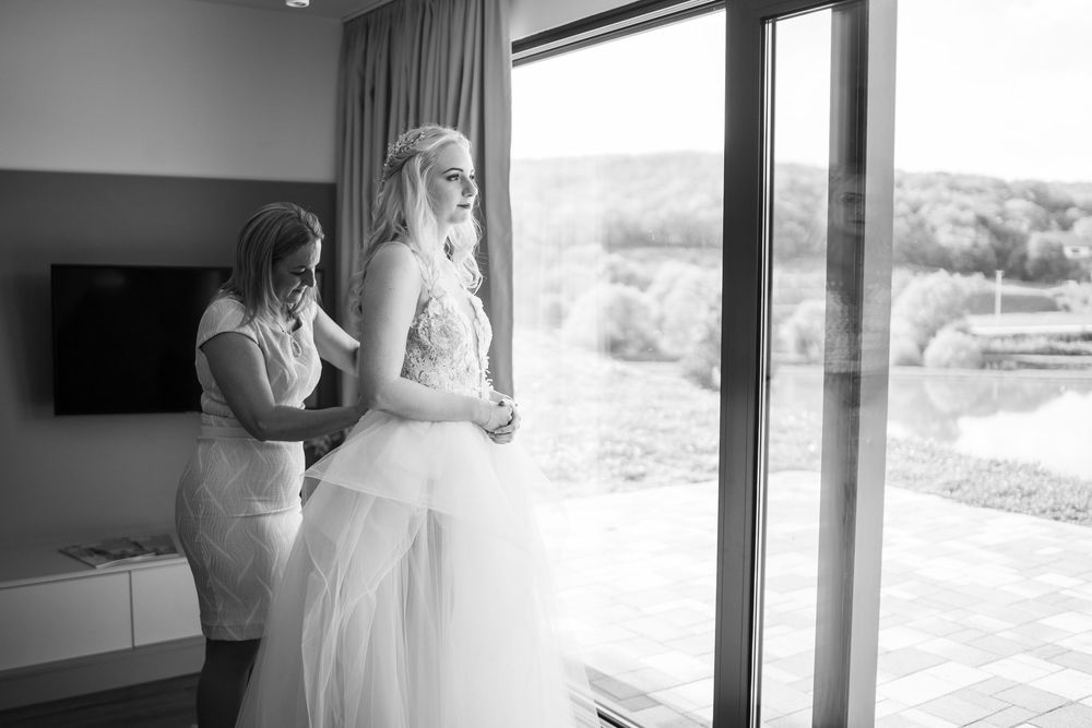 A wedding preparation scene showing a bridal gown being adjusted near large windows in a bright, modern room.