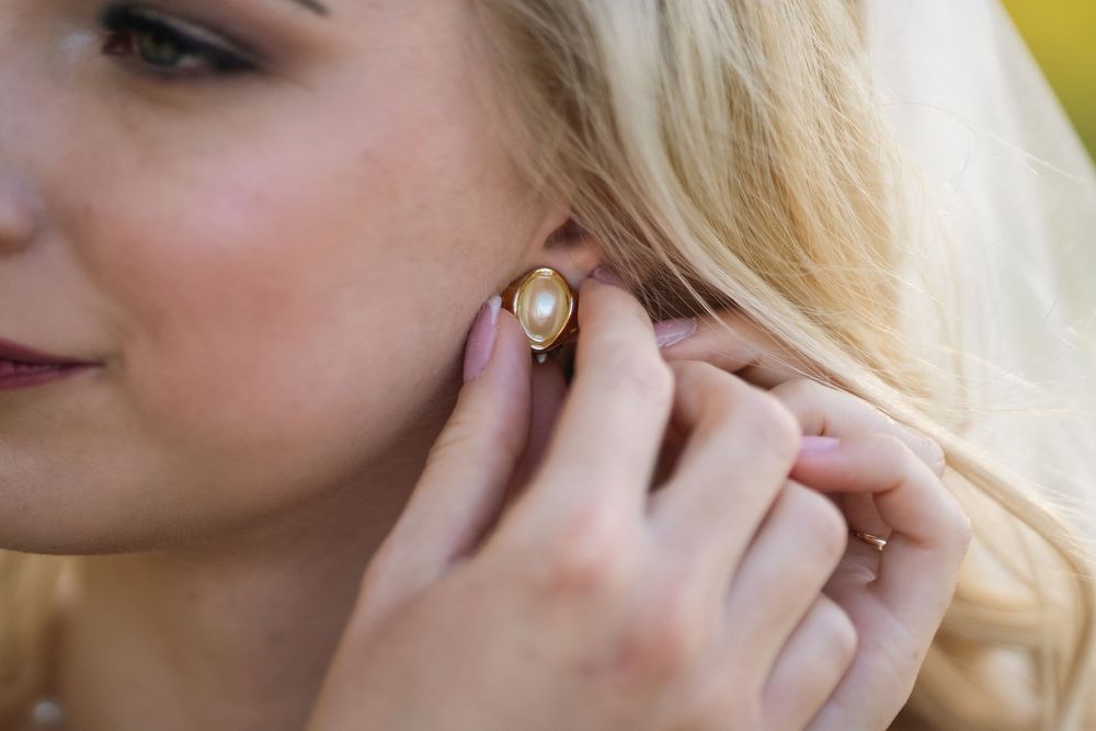 Close-up of hands putting on gold and pearl earring with blonde hair in background.