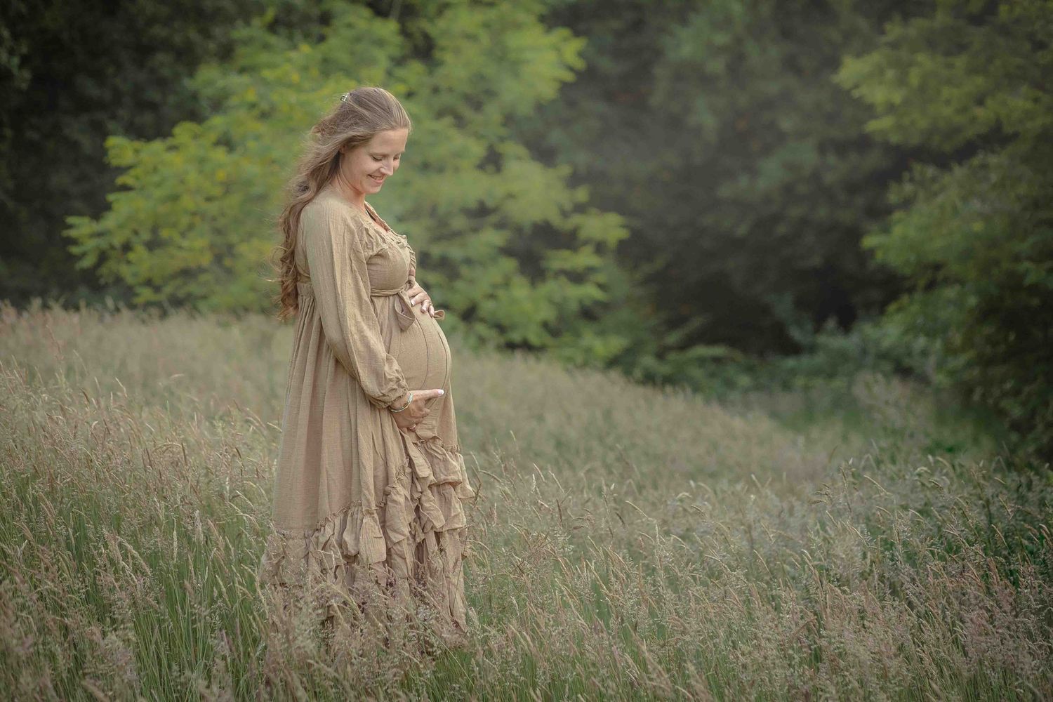 Zwangere vrouw in een lange jurk staat in een grasveld, handen op haar buik.