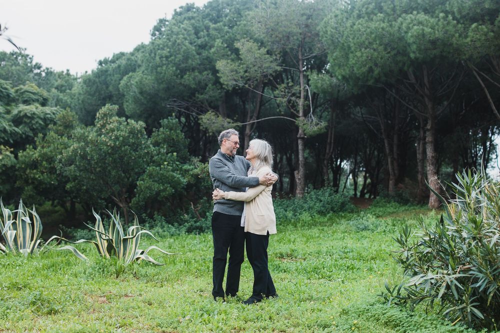 A couple embraces while standing in a lush green outdoor setting surrounded by trees and natural foliage.