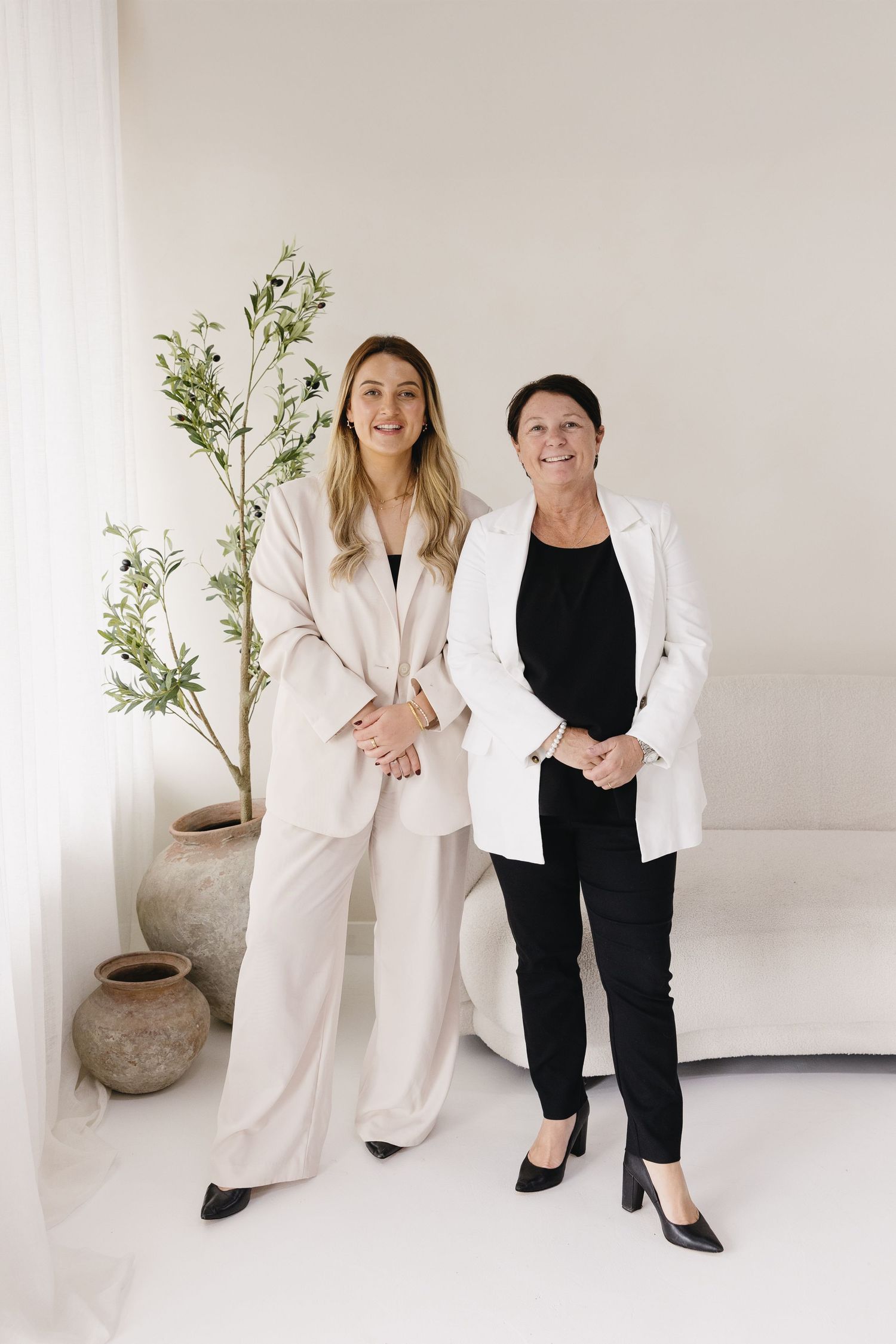 Two people in contrasting black and white outfits pose together in a minimalist studio setting with a potted plant.