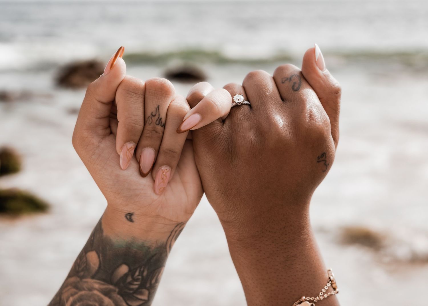 two people women with their pinky fingers linked together, engagement ring on display with the ocean in the background.