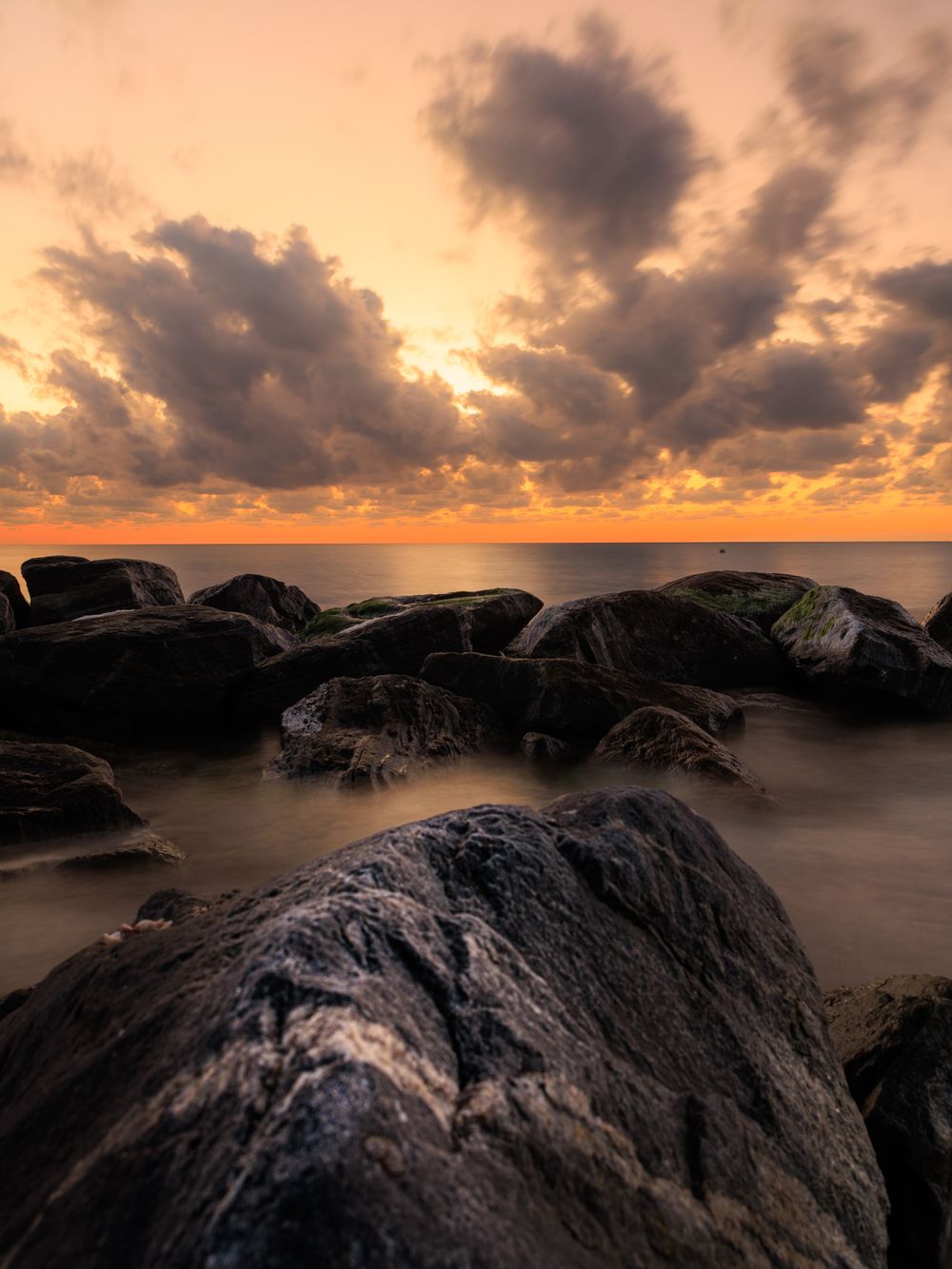 Rugged coastal rocks silhouetted against a warm orange sunset sky with dramatic cloud formations.