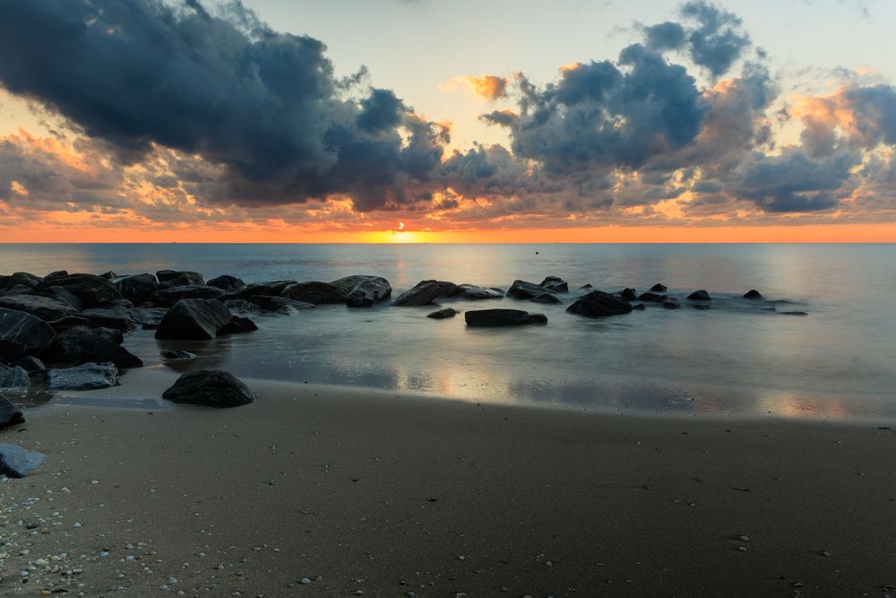 Dramatic seascape panorama with dark clouds and orange sunset reflecting on calm ocean waters.