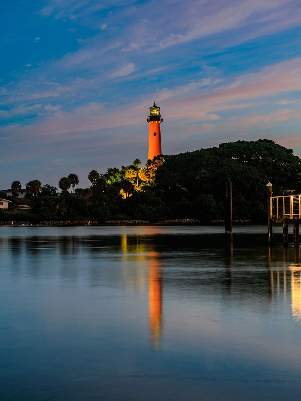 Red lighthouse reflecting on calm waters at dusk with vibrant orange glow.