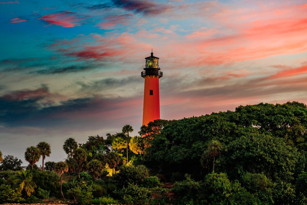 Scenic lighthouse silhouette against vibrant sunset sky with palm trees and rocky coastline.