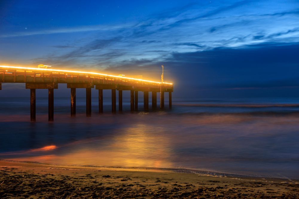 Illuminated pier at sunset reflecting golden light on the peaceful ocean surface.