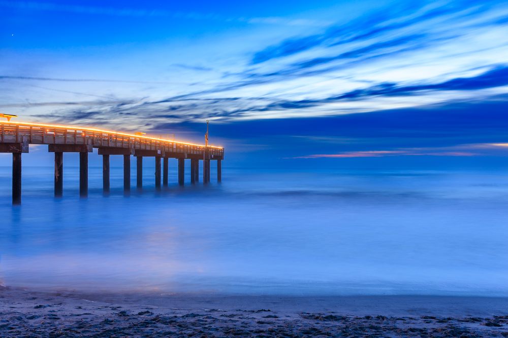 Long wooden pier extending into calm blue ocean waters during twilight.