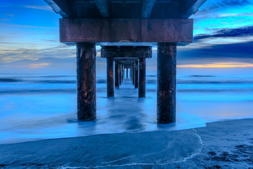 Underneath view of pier supports creating geometric patterns against blue water.