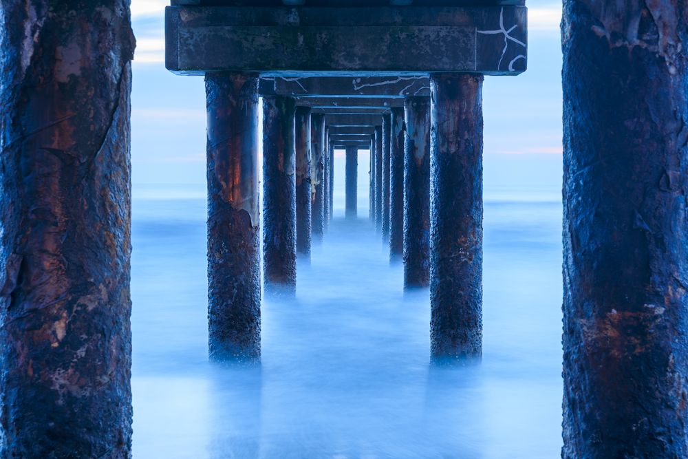 Dark wooden pier pillars silhouetted against smooth blue water during long exposure.