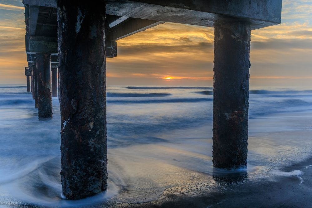 Long wooden pier extends into calm ocean waters during a dramatic golden sunset.