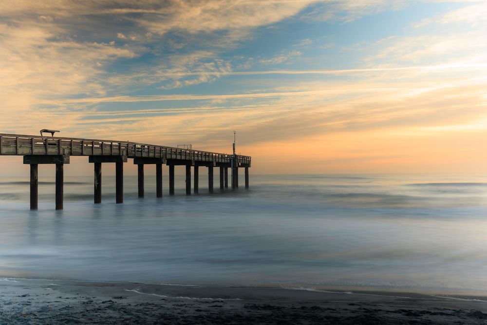 Panoramic view of a wooden pier stretching into the ocean at sunset with cloudy skies.