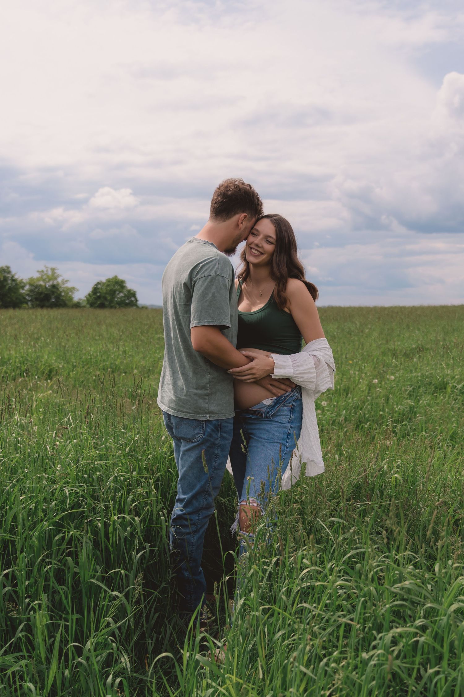A romantic couple embraces while standing in a tall grass field under cloudy skies.