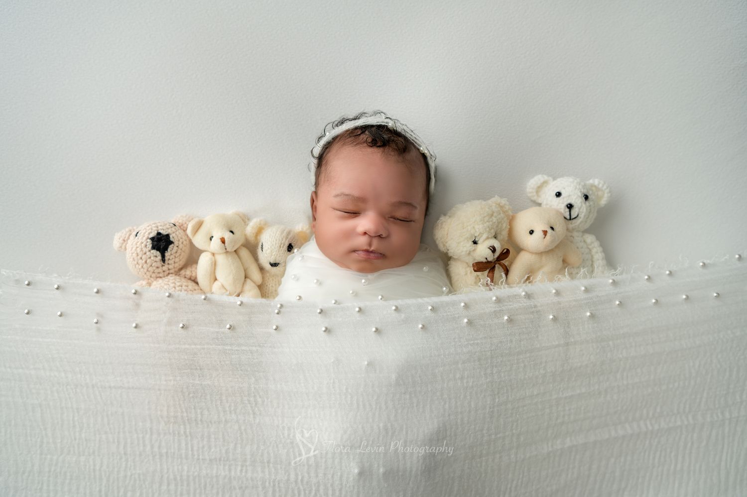 Sleeping baby nestled in white blanket with teddy bear stuffed animals lined up on both sides.