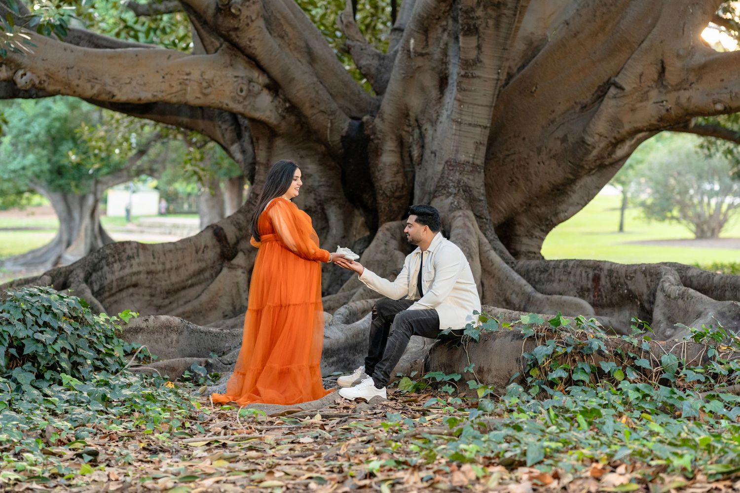Two people pose for engagement photos under a large tree canopy in a romantic outdoor setting.