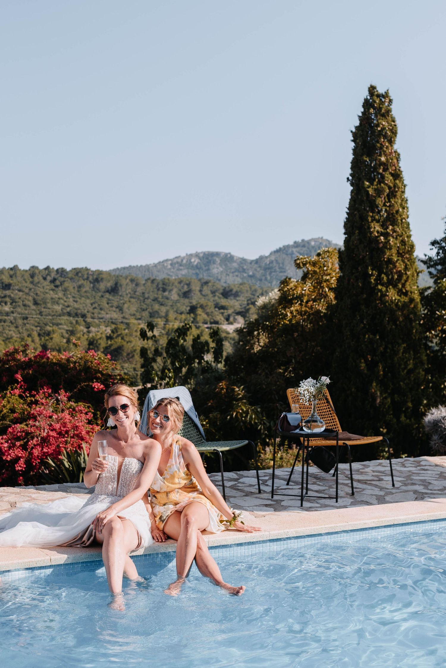 Zwei Freundinnen entspannen sich an einem Swimmingpool mit Blick auf die Berge und rosa Blumen in einer luxuriösen mediterranen Umgebung.
