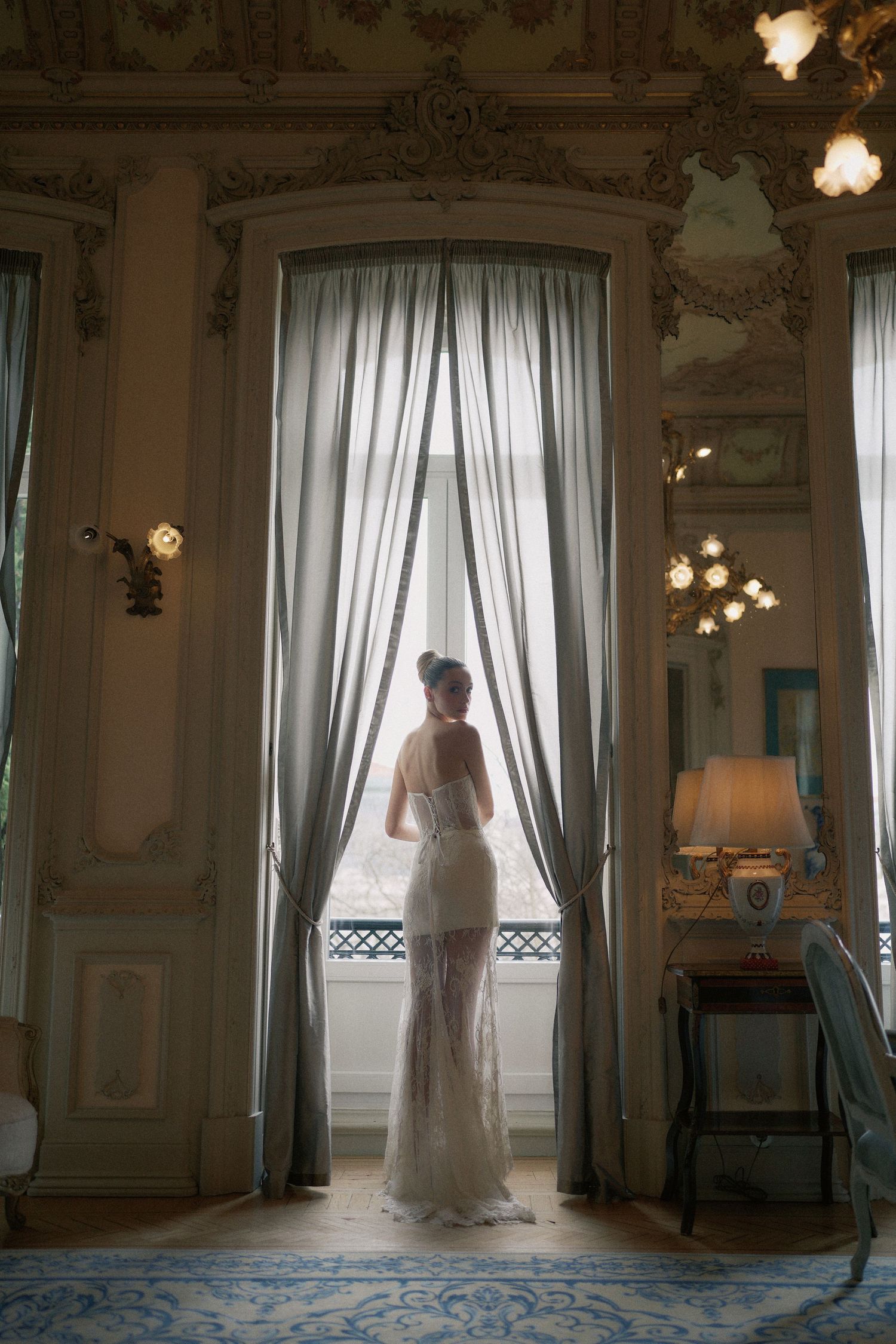 Elegant silhouette stands in sheer white dress by tall curtained window in ornate vintage room with crystal light fixtures.