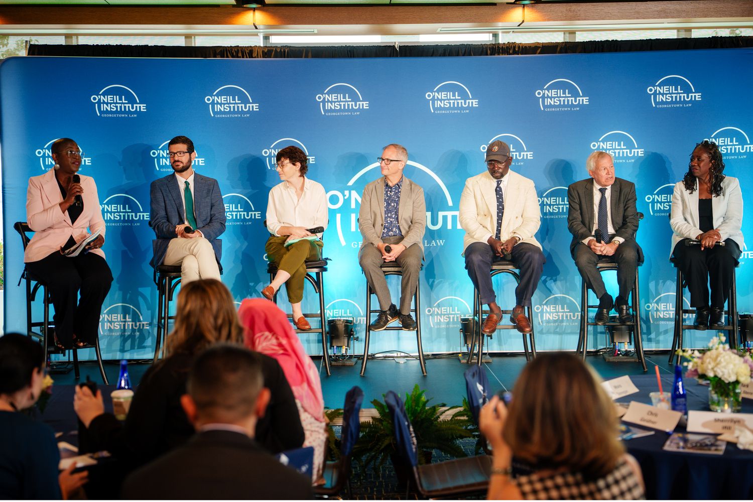 Business professionals seated at a conference panel table with blue backdrop discussing topics during a corporate event.