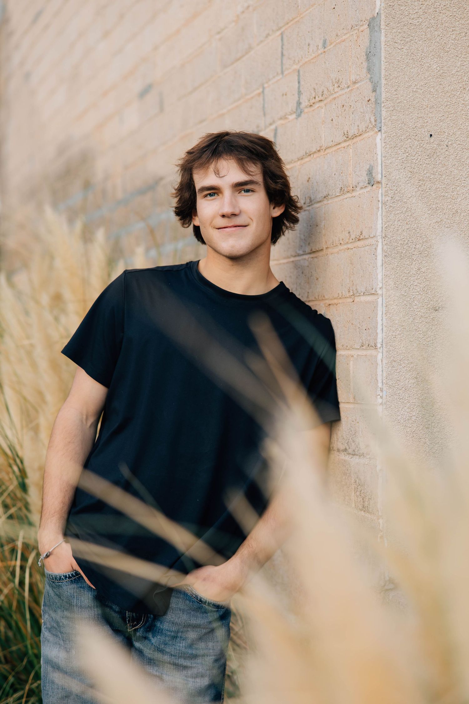 Young man in black t-shirt leaning against brick wall surrounded by tall golden grass.
