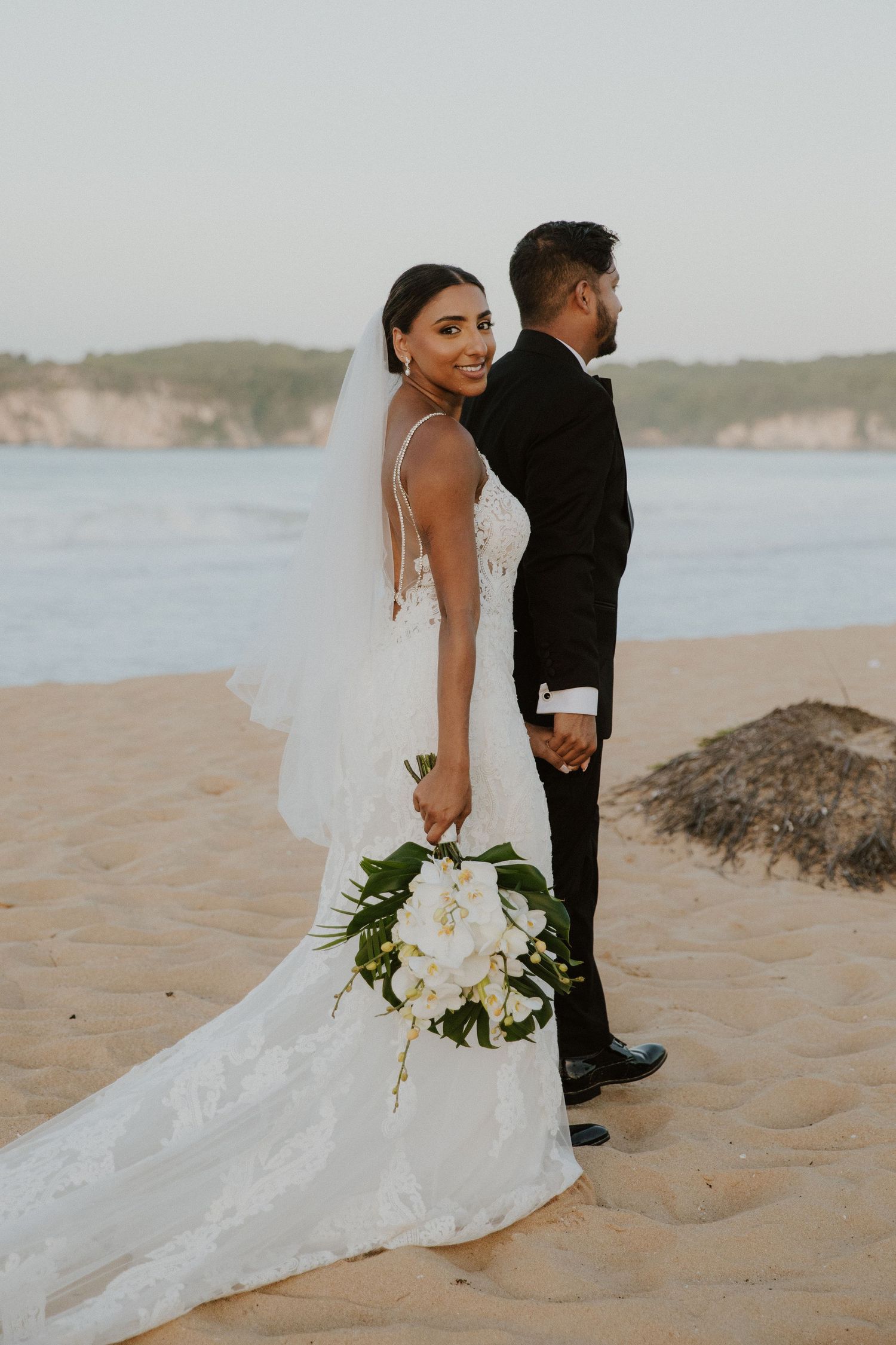 Wedding couple poses on beach with dramatic lace train and white floral bouquet.