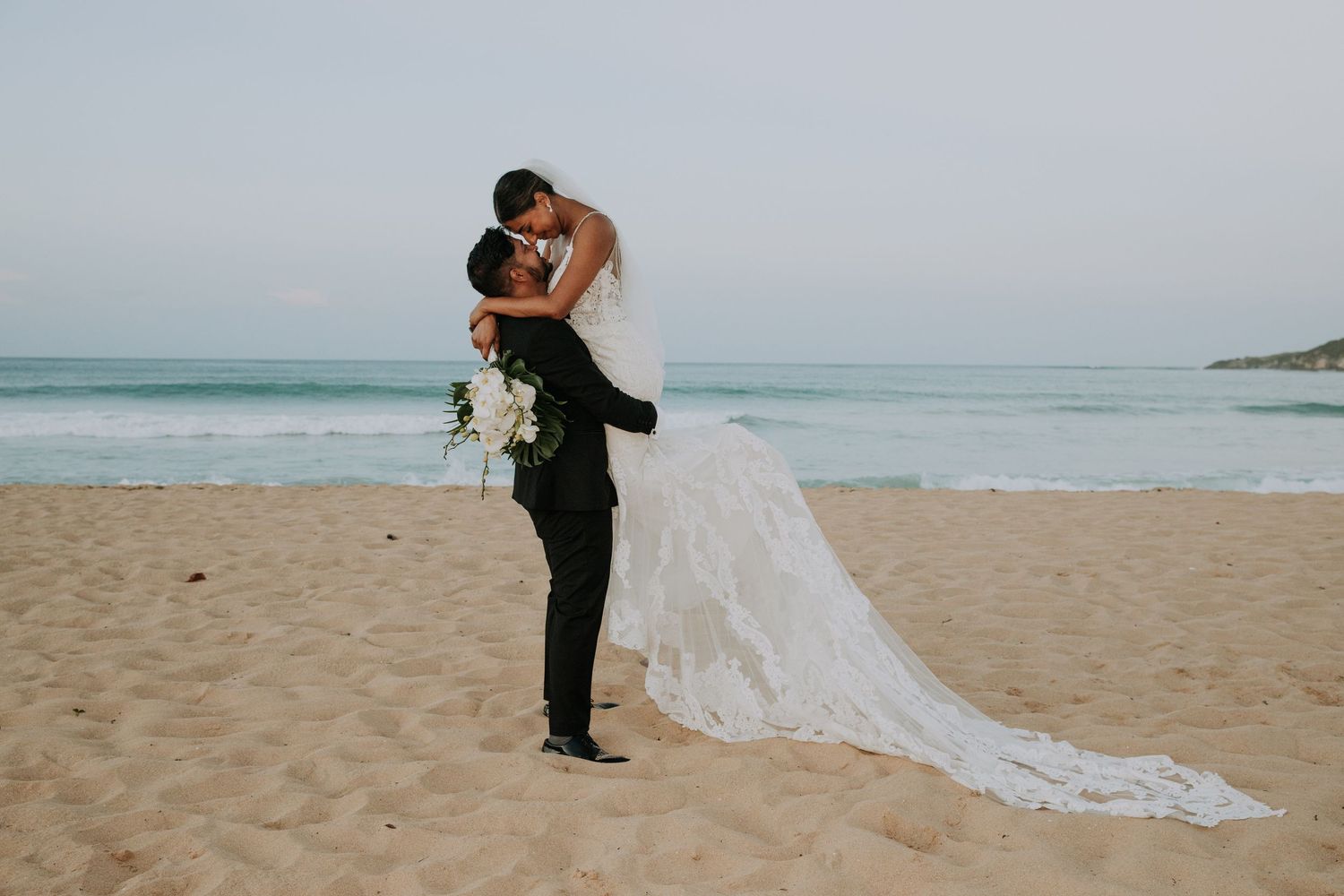 Groom lifts bride in an embrace on sandy beach with flowing white wedding dress train.
