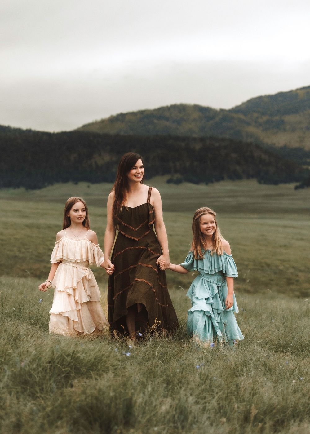 Mother walking with two daughters in ruffled dresses across a green meadow.