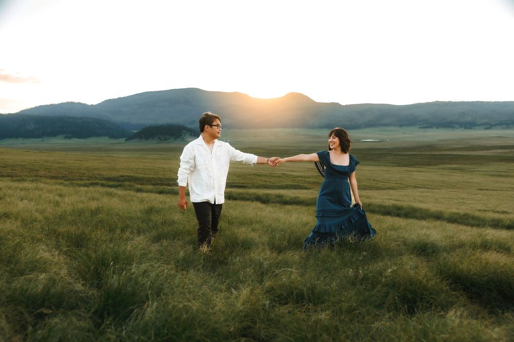 Couple holding hands walking through a green meadow with mountains at golden sunset.