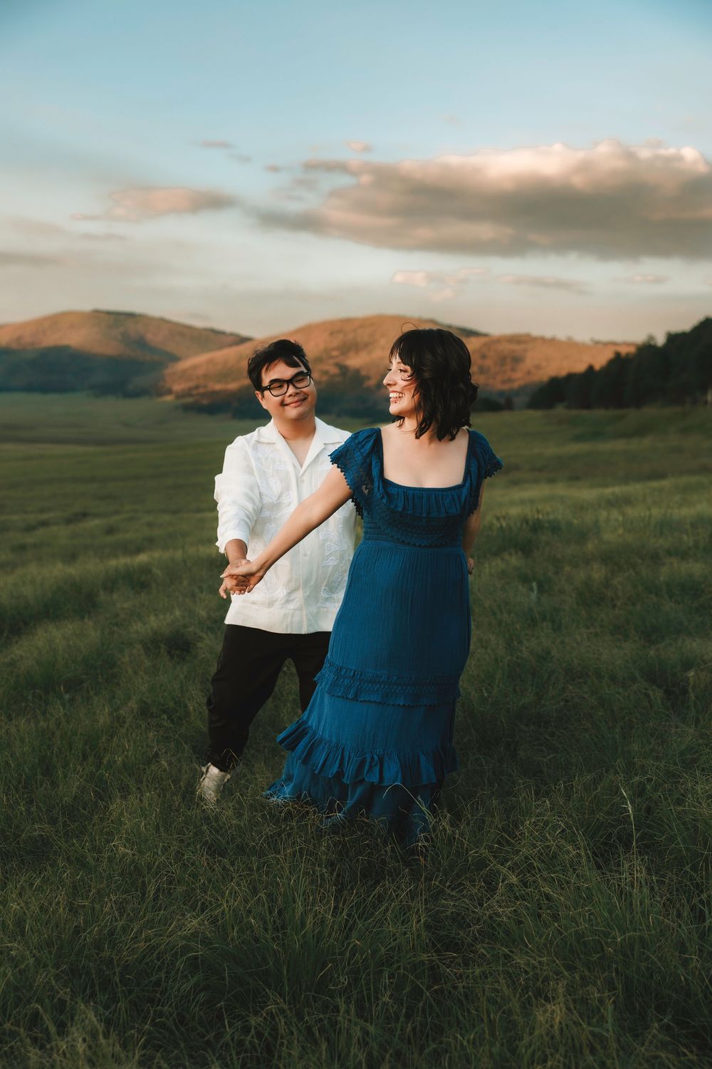 Smiling engaged couple holding hands in a green meadow with Jemez mountains and sunset sky.