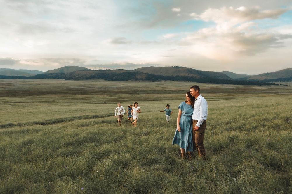Couple standing in a green meadow at sunset while children play in the background.