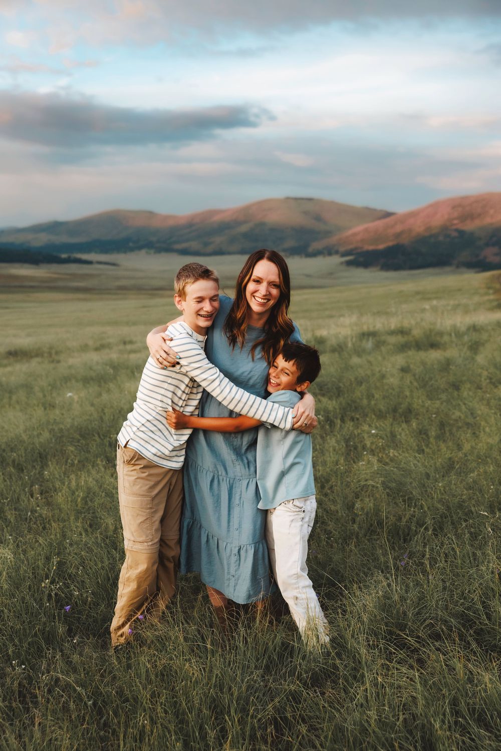 A smiling mother in a blue dress embraces her two sons in a sunlit green meadow with mountains.