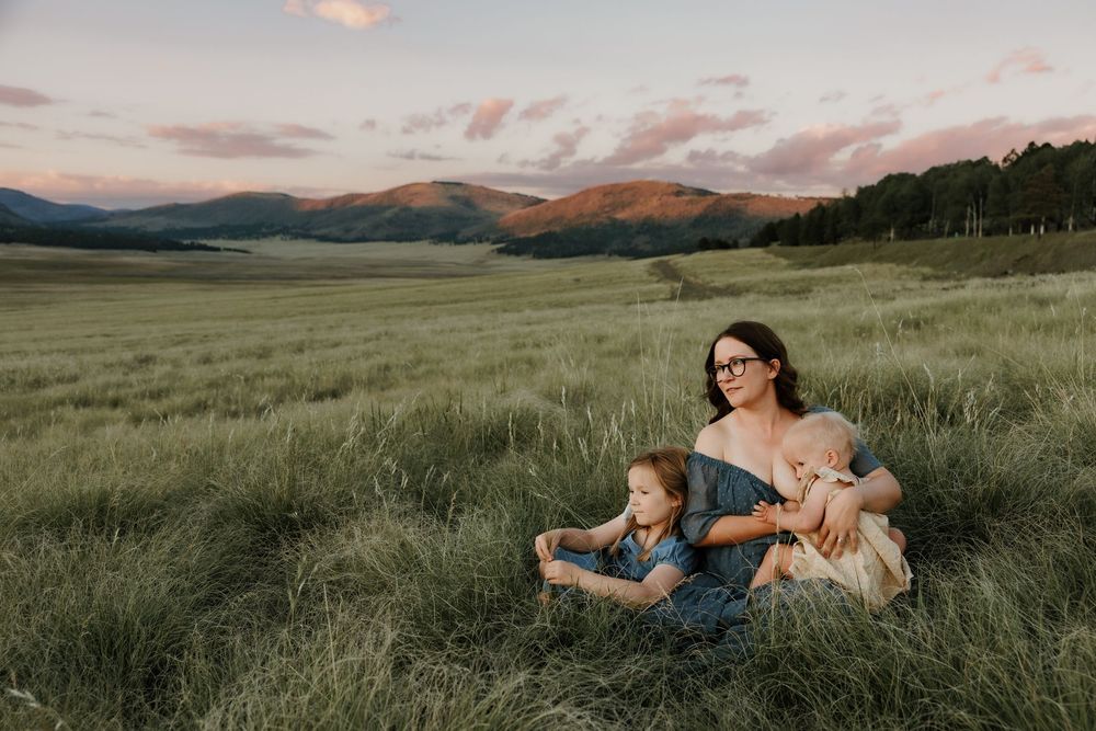 A mother sits in a lush green meadow with two young children at sunset, mountains visible in the background.