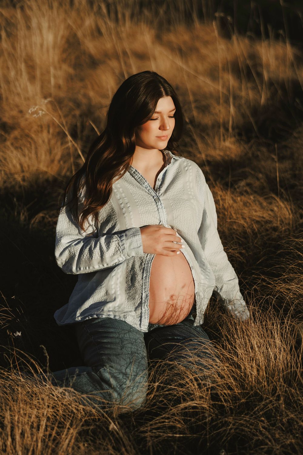 Pregnant woman sitting in golden grass field, cradling bare baby bump, wearing open denim shirt, bathed in warm sunlight.
