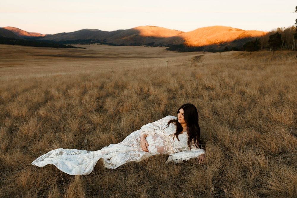 Woman in white floral dress lying in golden grass field with mountain backdrop at sunset.