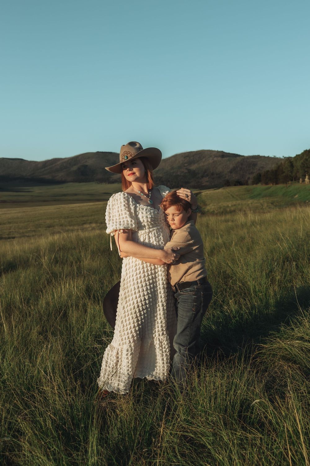 Mother in white dress and hat embracing her son in a sunlit grassy field.