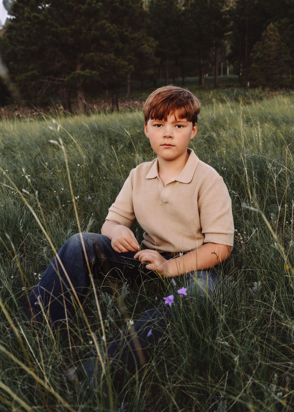 Young boy in beige polo shirt sitting in tall green grass with wildflowers.