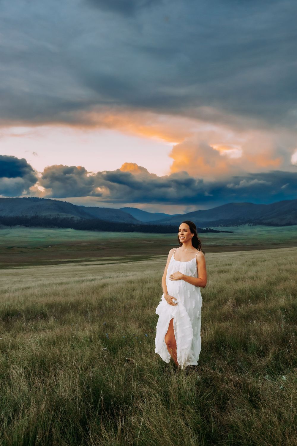 A pregnant woman in a flowing white dress stands in a green field under a dramatic sunset sky.