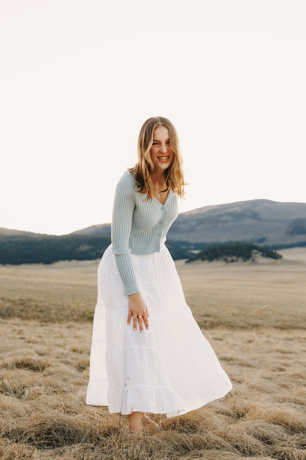 Smiling young woman in a light blue sweater and white maxi skirt standing in a golden field with mountains behind her.
