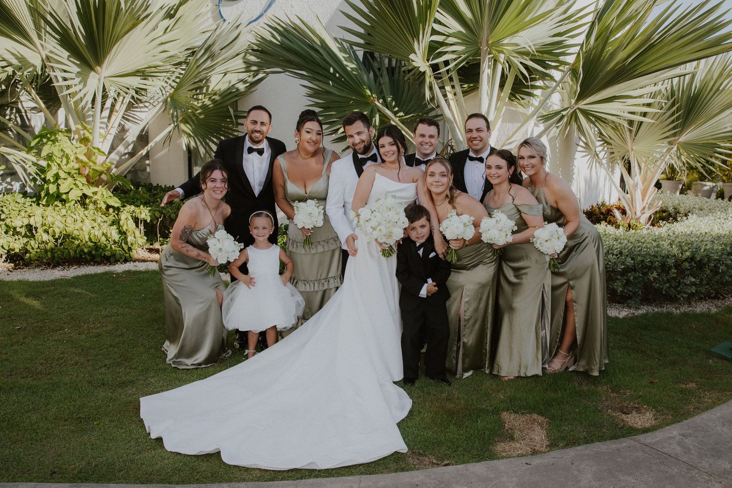 Wedding party poses together outdoors with palm trees and greenery as backdrop for formal group photo.