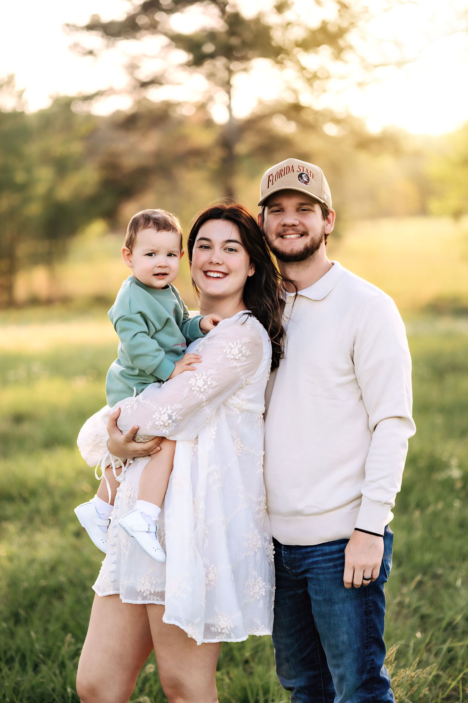 A young family poses together in a grassy field during golden hour sunset with warm backlighting.