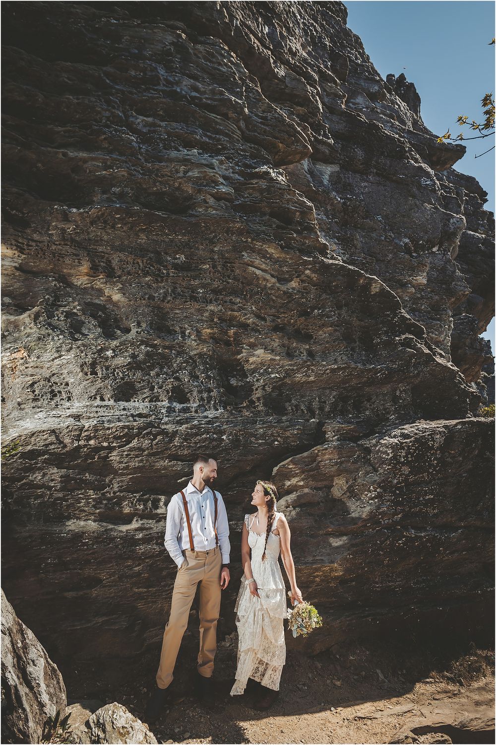A couple stands against dramatic rocky cliffs during their outdoor wedding photoshoot.