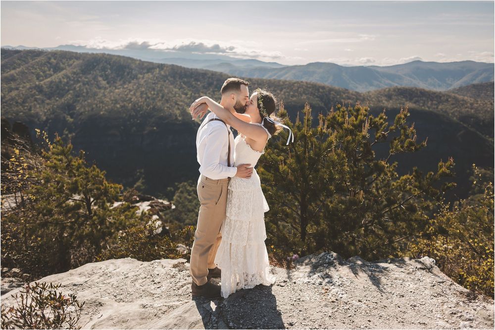 A couple embraces on a mountain peak overlooking a scenic mountain range landscape on a sunny day.