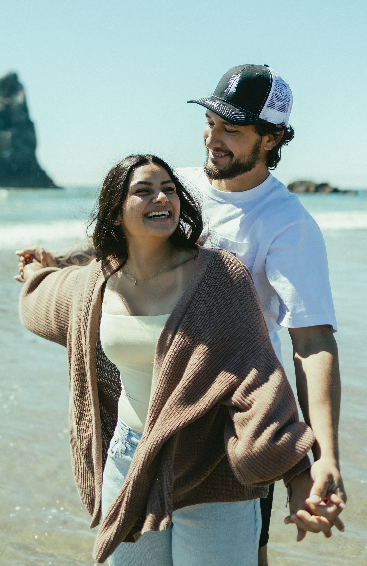 A couple shares a joyful moment together on a scenic beach with dramatic coastal rock formations in the background.
