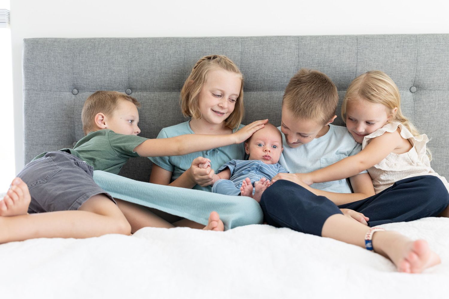 5 kids sitting on a bed with a newborn in the middle. all looking at him one boy touching babies head