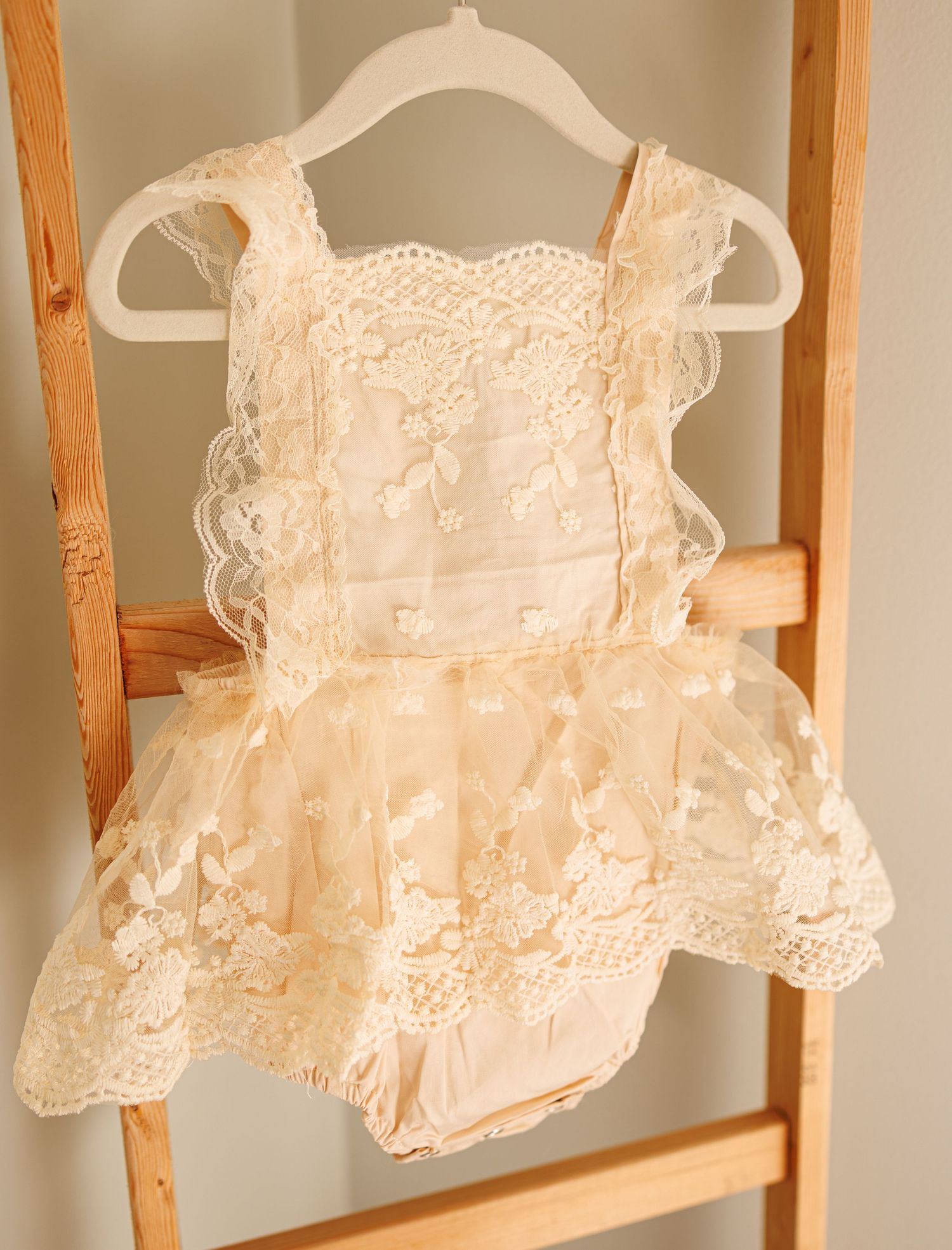 Delicate vintage-style cream lace baby romper with ruffles hanging on a white hanger against wooden ladder.