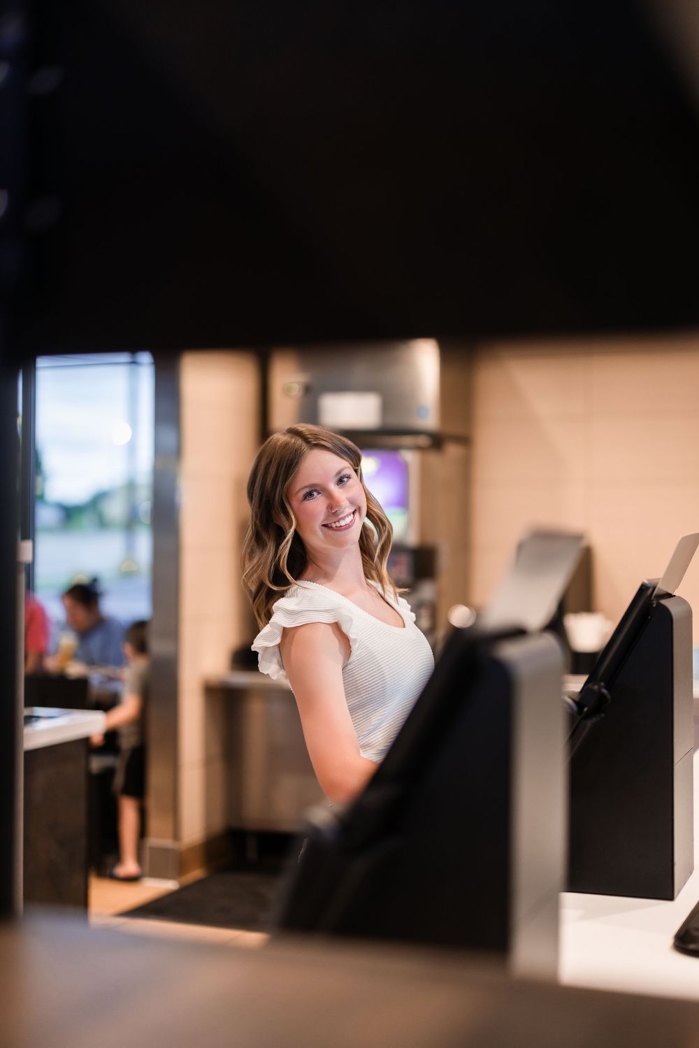 A woman wearing a white dress smiles while working at a business counter with blurred background decor.